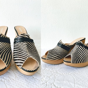 May include: A pair of open-toe platform mules with a zebra print pattern. The shoes have a black leather strap across the top and a wooden heel. The platform sole is also made of wood.