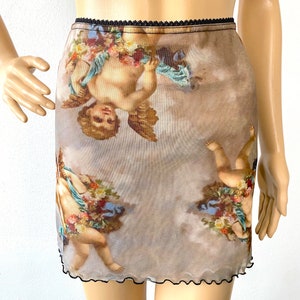 Puede incluir: Una minifalda transparente de tul con cintura el&aacute;stica negra y dobladillo festoneado negro. La falda presenta un estampado de querubines con flores sobre un fondo nublado.