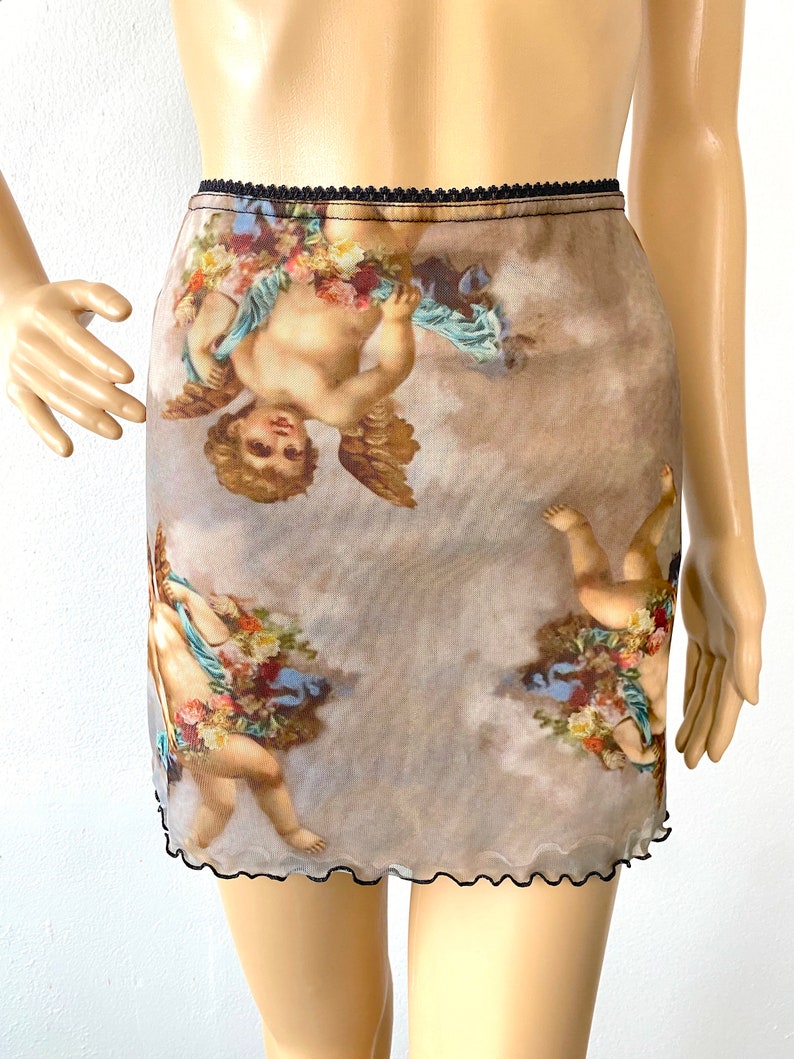 Puede incluir: Una minifalda transparente de tul con cintura el&aacute;stica negra y dobladillo festoneado negro. La falda presenta un estampado de querubines con flores sobre un fondo nublado.