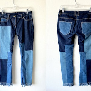 Op de afbeelding: Donkerblauwe denim jeans met lichtblauwe patchwork op de voor- en achterkant. De jeans heeft een klassiek ontwerp met vijf zakken en een gerafelde zoom. De jeans hangt aan een doorzichtige hanger tegen een witte achtergrond.