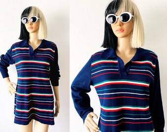 May include: A navy blue long-sleeve sweater dress with horizontal stripes in red, white, and green. The dress has a collared neckline and a relaxed fit. The model is wearing white sunglasses.