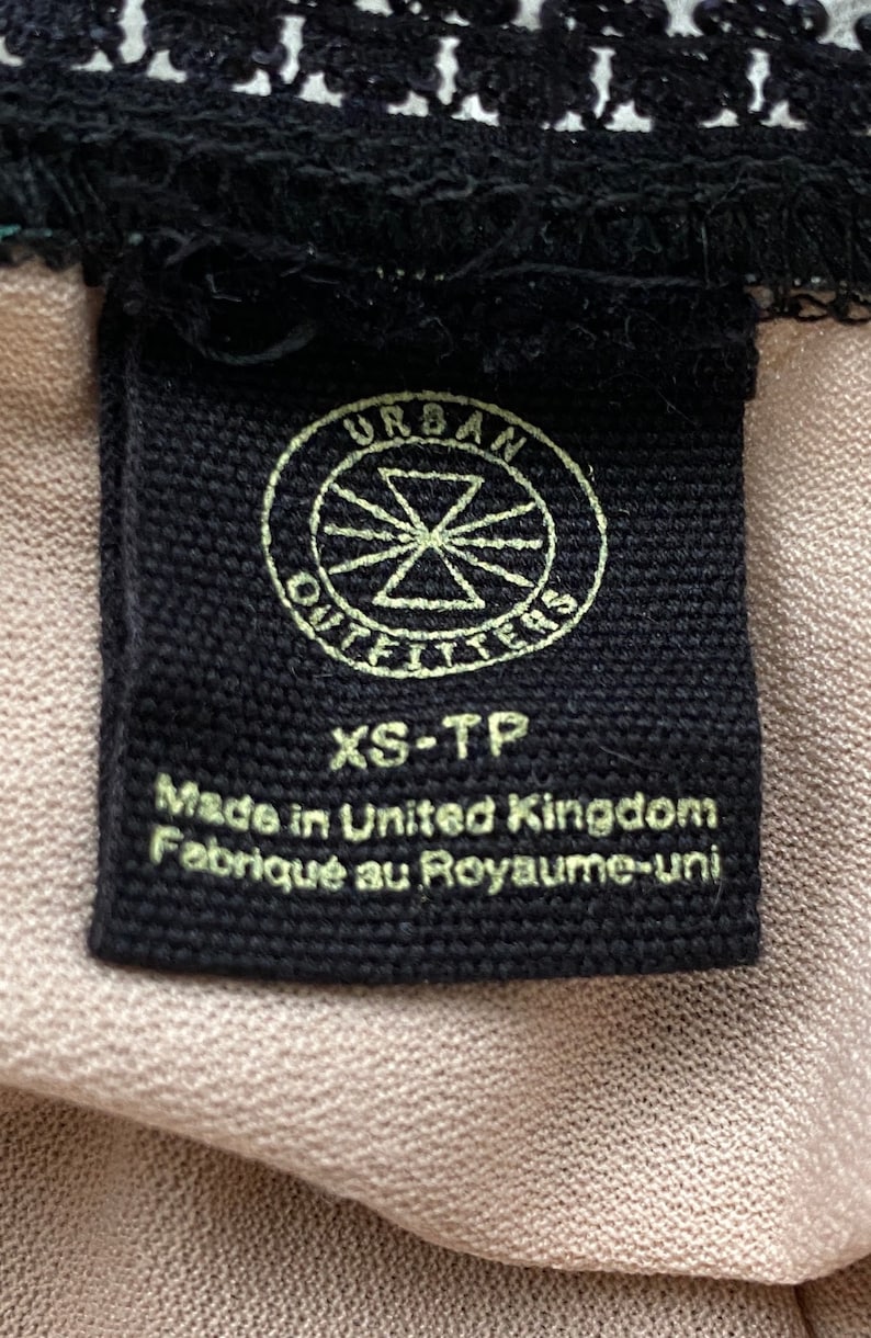 Puede incluir: Etiqueta de ropa negra con texto blanco que dice "Urban Outfitters" en un c&iacute;rculo con una estrella en el centro. Debajo del c&iacute;rculo, el texto dice "XS-TP" y "Made in United Kingdom Fabriqu&eacute; au Royaume-Uni."