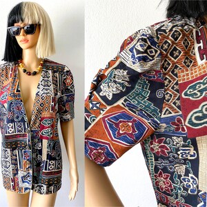 Peut inclure: Une veste à manches courtes, à ouverture sur le devant, avec un motif géométrique coloré. Le tissu présente un mélange de rouge, d'orange, de bleu, de vert et de blanc. La veste a une fermeture à bouton et un col.