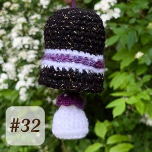 Può includere: Una campana all'uncinetto fatta a mano con una parte superiore nera, impreziosita da fili d'oro, e una striscia bianca e viola. La campana ha una base bianca e una parte superiore viola. L'immagine include il testo "#32" in un riquadro bianco.