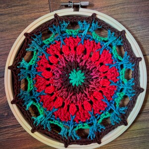 May include: A colorful crocheted mandala with a red, brown, green, and blue color scheme. The mandala is framed in a wooden hoop.