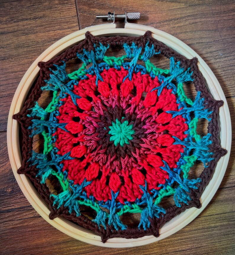 May include: A colorful crocheted mandala with a red, brown, green, and blue color scheme. The mandala is framed in a wooden hoop.