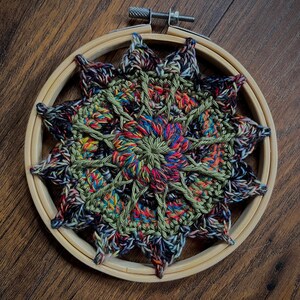 May include: A colorful crocheted mandala with a green, red, blue, and black color scheme is displayed in a wooden embroidery hoop.