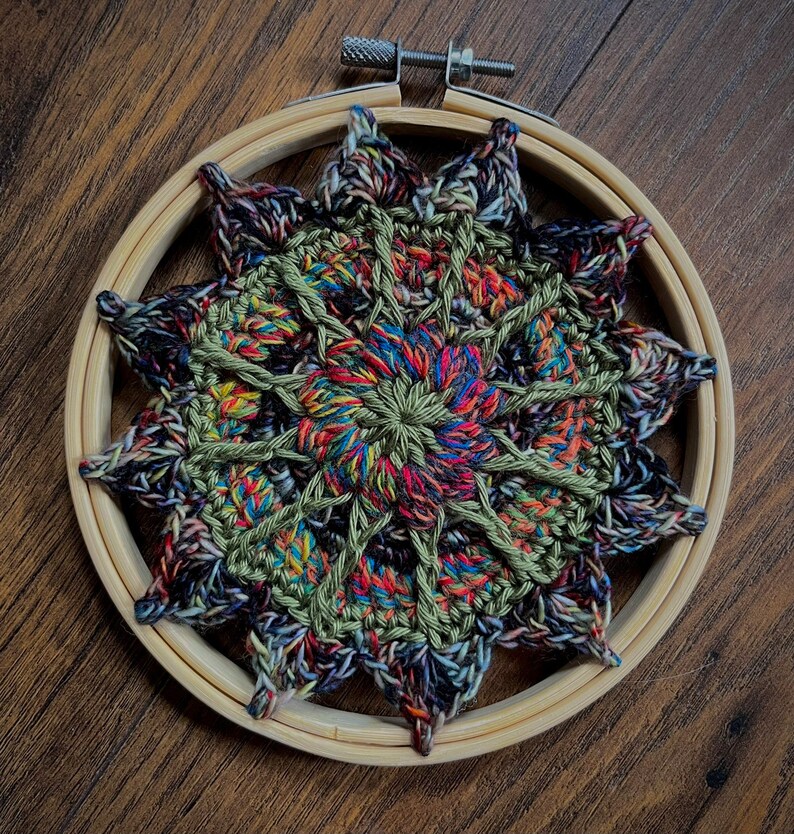 May include: A colorful crocheted mandala with a green, red, blue, and black color scheme is displayed in a wooden embroidery hoop.