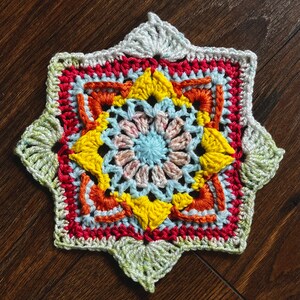 May include: A crocheted star-shaped doily with a multi-colored design. The center of the doily is a light blue and pink flower with yellow petals. The doily is surrounded by a red, orange, and yellow border.
