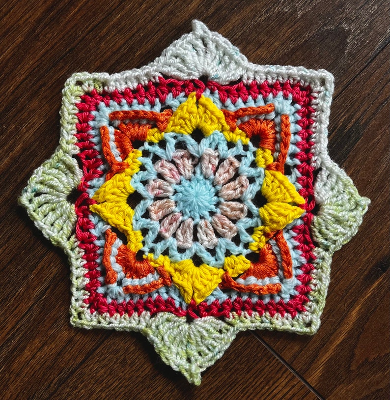May include: A crocheted star-shaped doily with a multi-colored design. The center of the doily is a light blue and pink flower with yellow petals. The doily is surrounded by a red, orange, and yellow border.