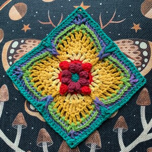 May include: A crocheted square with a flower design in the center. The flower is made with red, maroon, and yellow yarn. The square is bordered with teal, green, and purple yarn.