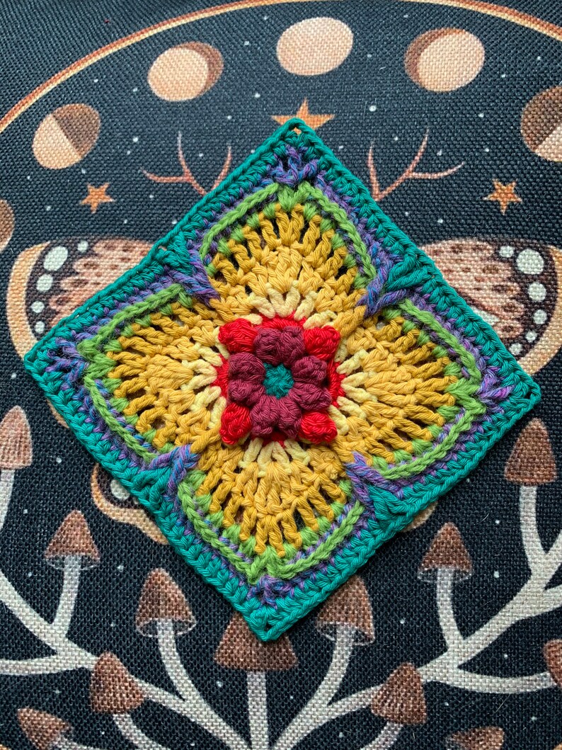 May include: A crocheted square with a flower design in the center. The flower is made with red, maroon, and yellow yarn. The square is bordered with teal, green, and purple yarn.