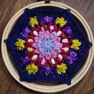 May include: A colorful crocheted mandala with a blue, purple, pink, red, yellow, and white color scheme. The mandala is framed in a wooden hoop.