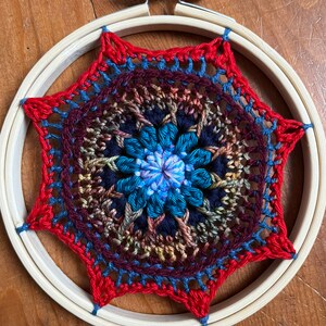 May include: A colorful crocheted mandala with a blue, teal, and purple center, surrounded by red, brown, and blue yarn. The mandala is framed in a wooden embroidery hoop.