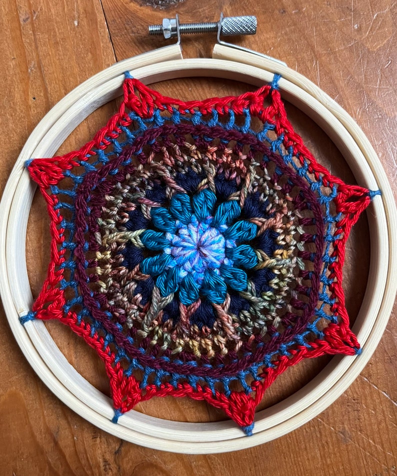 May include: A colorful crocheted mandala with a blue, teal, and purple center, surrounded by red, brown, and blue yarn. The mandala is framed in a wooden embroidery hoop.