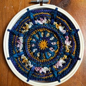 May include: A circular crocheted mandala with a blue, purple, gold, and pink color scheme. The mandala is framed in a wooden embroidery hoop.