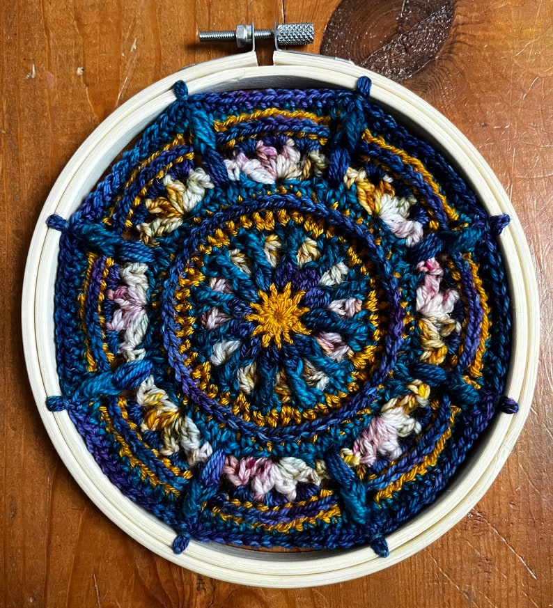 May include: A circular crocheted mandala with a blue, purple, gold, and pink color scheme. The mandala is framed in a wooden embroidery hoop.