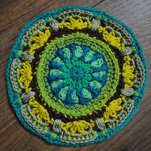 May include: A circular crocheted coaster with a multi-colored design. The center features a blue and green floral pattern surrounded by a brown, yellow, and green border.