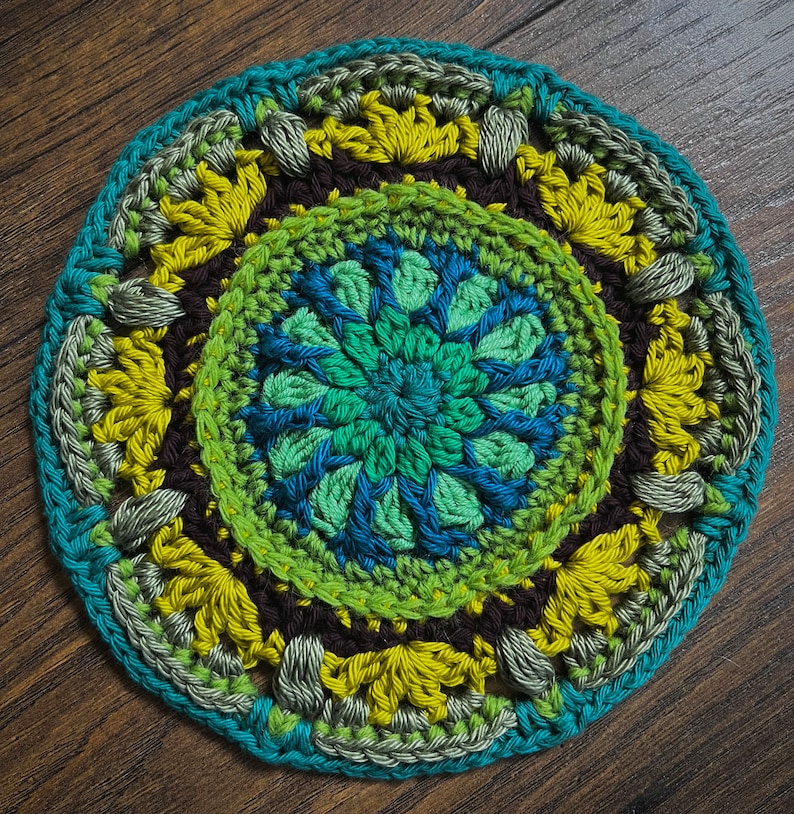 May include: A circular crocheted coaster with a multi-colored design. The center features a blue and green floral pattern surrounded by a brown, yellow, and green border.