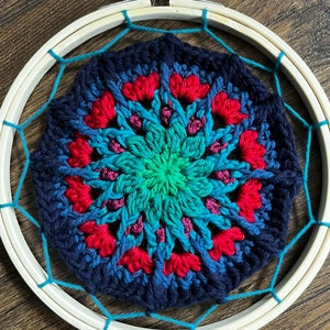 May include: A crocheted mandala with a blue, green, and red color gradient, framed in a wooden hoop with a turquoise string.