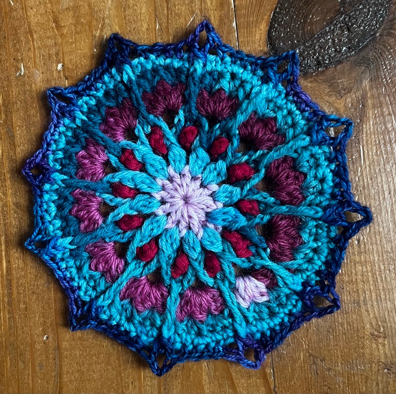 May include: A crocheted mandala with a blue, purple, and red color scheme. The mandala has a circular design with a central flower and intricate patterns radiating outwards.