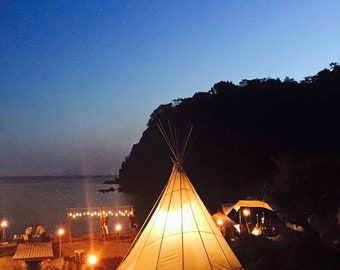 May include: A large white teepee tent illuminated from within, set up on a beach at night. The tent is surrounded by string lights and small lanterns. A dark silhouette of a cliff is visible in the background.