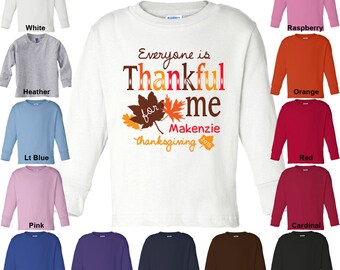 May include: White long-sleeve toddler t-shirt with the text "Everyone is Thankful for me Makenzie Thanksgiving 2015" and a graphic of fall leaves. The shirt is available in 12 colors: white, heather, light blue, pink, royal blue, purple, navy, brown, black, raspberry, orange, red, and cardinal.