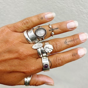 May include: A collection of silver rings of various designs. Rings include a ring with a large oval-shaped, light-colored stone, a ring with a small purple stone, and rings with etched patterns. The rings are worn on multiple fingers.