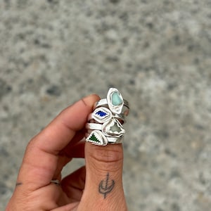 May include: Four silver rings with different colored glass stones. The rings are stacked on a person's thumb.