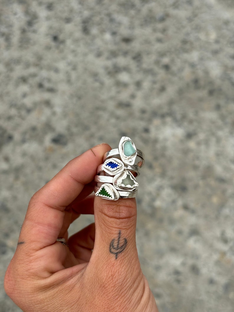 May include: Four silver rings with different colored glass stones. The rings are stacked on a person's thumb.