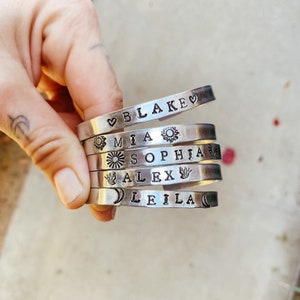 May include: A stack of six silver bracelets with different names and symbols engraved on them. The names are Blake, Mia, Sophia, Alex, and Leila. The symbols include a heart, a sun, a flower, a bird, and a crescent moon.