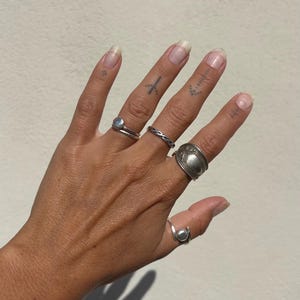 May include: A hand wearing several silver rings. The rings include a double band ring with a small stone, a twisted band, a large oval ring, and a ring with a small sphere. The hand has several small tattoos.