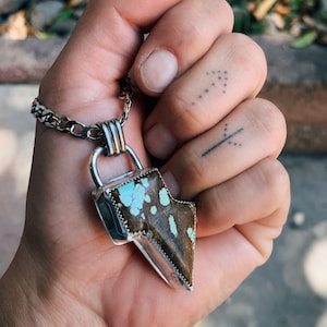 Puede incluir: Un collar con colgante de plata con una piedra turquesa y marrón en forma de rayo. El colgante está sujeto a una cadena de plata. El collar se sostiene en una mano.