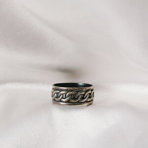 May include: A silver ring with a braided design. The ring is on a white fabric background.