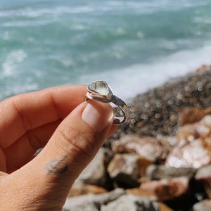 May include: A silver ring with a clear, triangular sea glass stone set in the band. The ring is being held by a hand against a background of rocks and ocean waves.