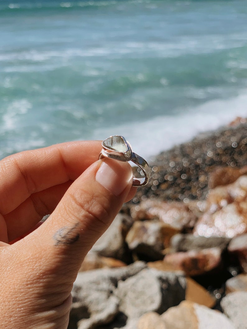 May include: A silver ring with a clear, triangular sea glass stone set in the band. The ring is being held by a hand against a background of rocks and ocean waves.