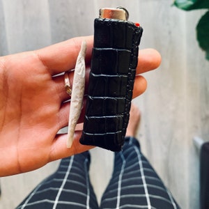 Pu&ograve; includere: Un porta sigarette in pelle nera con un motivo a coccodrillo e un accendino argentato. Il porta sigarette &egrave; tenuto in una mano.