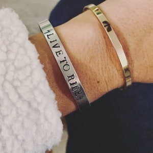 May include: A silver bracelet with the words "Live to Ride" engraved on it. The bracelet is worn on a person's wrist with a gold bracelet on top of it.