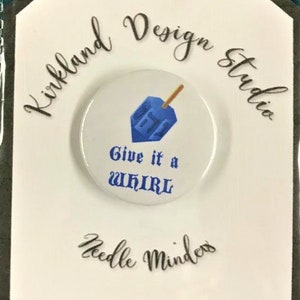 May include: A white tag with black text that reads "Kirkland Design Studio" and "Needle Minder". The tag also has a blue and white button with a blue dreidel and the text "Give it a whirl".