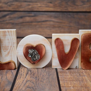 May include: Wooden block letters spelling "LOVE" with a heart-shaped cutout in the "O" block featuring a dog silhouette. The letters are painted in a reddish-brown color and have a white background.