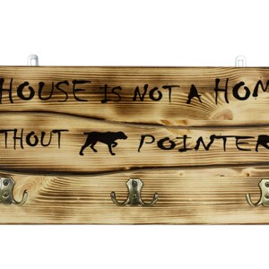 May include: A wooden sign with three hooks reads "A house is not a home without a pointer" with a silhouette of a dog. The sign has a rustic, burnt wood finish.