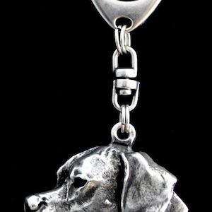 Può includere: Un portachiavi color argento con un ciondolo a forma di testa di cane dettagliata. Il ciondolo, con un profilo di cane strutturato, è attaccato a un moschettone e una catena. Lo sfondo è nero.