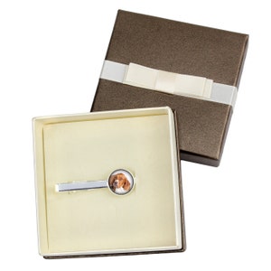 May include: A silver tie clip with a round, white, and silver-rimmed photo insert featuring a brown and white dog. The tie clip is in a brown gift box with a white satin ribbon.