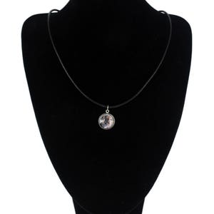 Puede incluir: Un collar gargantilla negro con un colgante plateado que presenta un cabujón redondo de vidrio transparente con una imagen en blanco y negro en su interior. El collar se muestra en un busto de terciopelo negro.