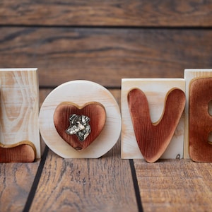 Puede incluir: Letras de madera que deletrean "LOVE" con un bloque en forma de corazón en el medio que presenta una pequeña figura de perro. Las letras están pintadas de color marrón rojizo y el corazón es de un rojo más oscuro.