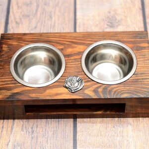 May include: A wooden pet bowl holder with two stainless steel bowls. The dark brown stained wood has a decorative metal rose in the centre. The bowls are round and shiny, ideal for feeding pets.