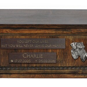 May include: A wooden pet urn with a brown finish. It has a plaque with the inscription "YOU LEFT OUR LIVES, BUT YOU WILL NEVER LEAVE OUR HEARTS" and a second plaque with the name "CHARLIE" and the dates "17.07.2005 † 25.02.2018". A metal dog head is attached to the urn.