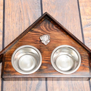May include: A wooden dog bowl stand in the shape of a house with two stainless steel bowls. The stand has a dark brown wood finish and a metal dog head ornament on the front.