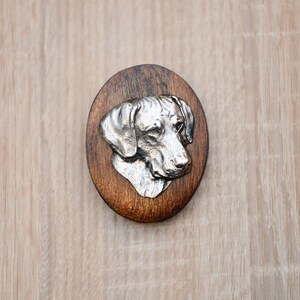May include: A silver-colored dog head sculpture mounted on a round wooden plaque. The dog has a short, smooth coat and is looking to the side.