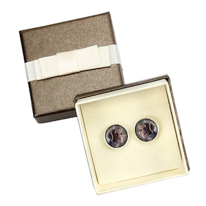 May include: A pair of brown and white dog portrait stud earrings in a cream-colored gift box with a brown lid and a white satin bow.